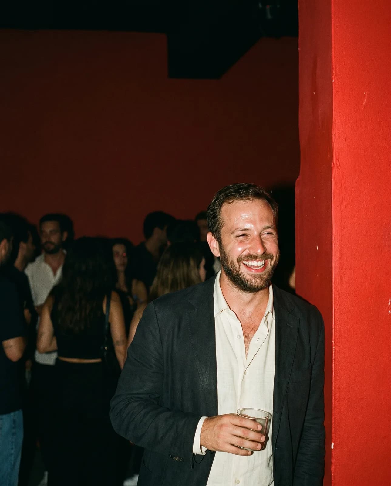 Campaign image: a man laughing, drink in hand, leaning on a red pillar